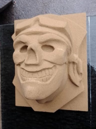 Reliefs des Gesichts eines Grimassen schneidenden Schädels auf einem quadratischen Hintergrund.