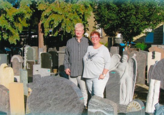 Paar steht zwischen Grabsteinen auf einem Friedhof. Grüne Bäume im Hintergrund.
