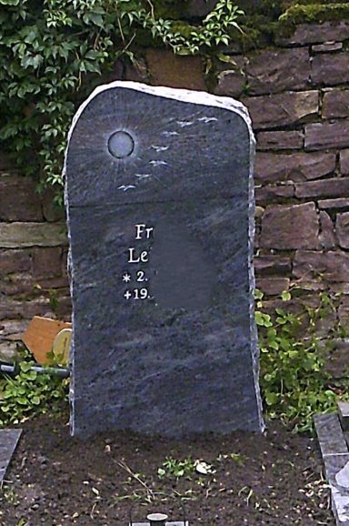 Schwarzer Grabstein mit Inschriften und umgebender Vegetation.