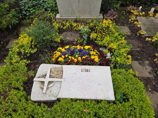 Grabstein mit Kreuz, umgeben von bunten Blumen und grünem Bodendecker.