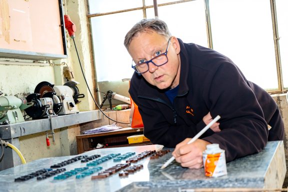 Mann arbeitet konzentriert an einem Tisch, malt mit Pinsel und Farbbehälter.