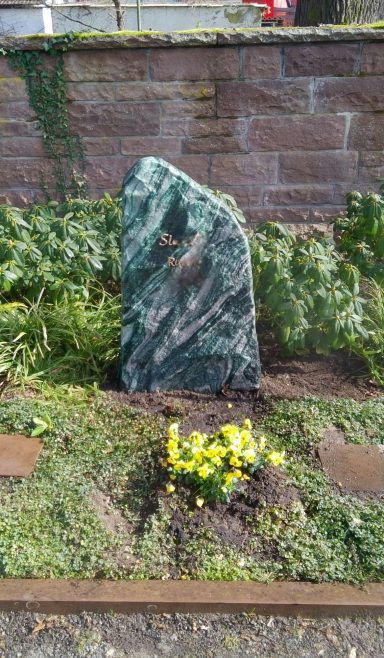 Grüner Findling umgeben von Pflanzen und gelben Blumen in einem Garten.