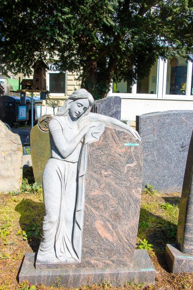 Grabstein mit einer trauernden, geflügelten Figur, umgeben von anderen Grabsteinen.