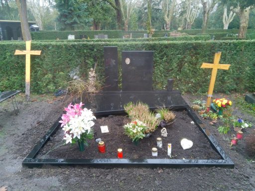 Grabstelle mit Blumen, Kerzen und Holzkreuzen, umgeben von einer Hecke.
