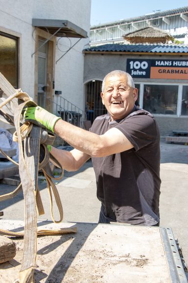 Mann arbeitet mit Werkzeugen auf einer Baustelle, lächelt in die Kamera.