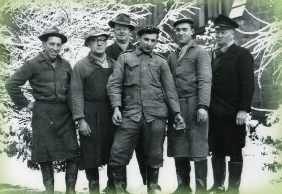 Sechs Männer in historischen Outfits stehen lächelnd vor einer schneebedeckten Kulisse.
