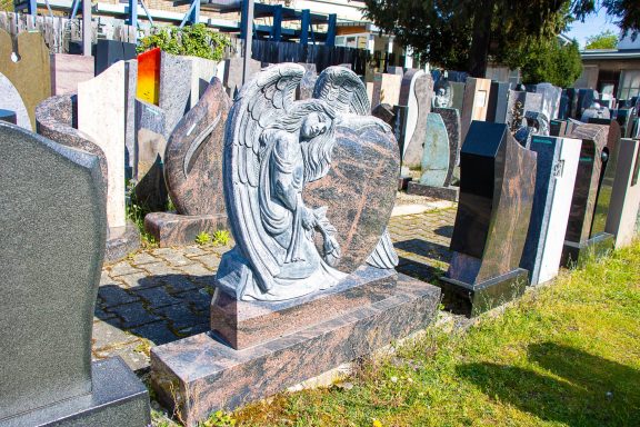 Gravestone mit Engel und Schild auf einem Friedhof, umgeben von weiteren Grabstätten.