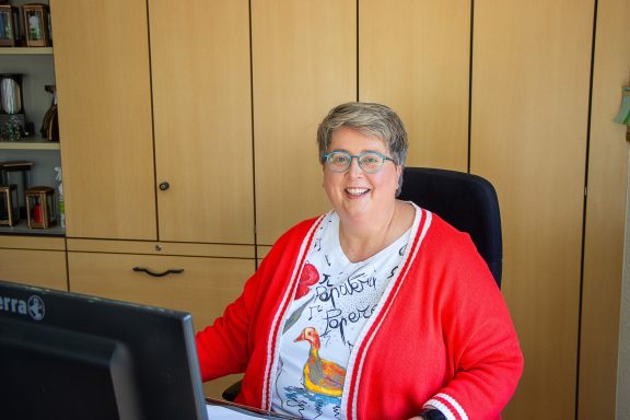 Lächelnde Frau mit Brille in roter Strickjacke vor einem Computer in einem hellen Raum.