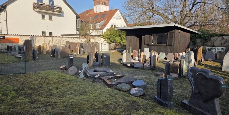 Friedhof mit Grabsteinen und einer Holzhütte im Hintergrund bei sonnigem Wetter.