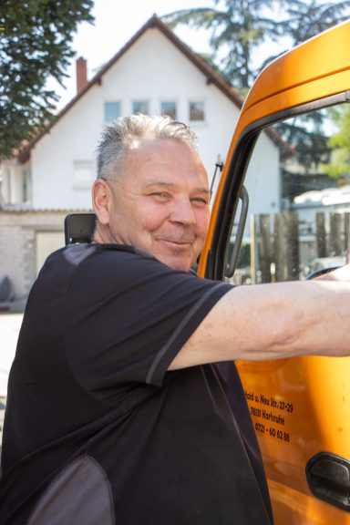 Lächelnder Mann neben einem orangefarbenen Fahrzeug, im Hintergrund ein Haus.