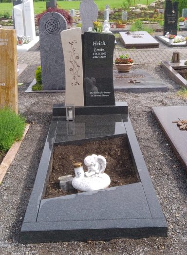 Grabstein mit zwei Steinen, einer schwarz und einer beige, umgeben von Rasen und Blumen.