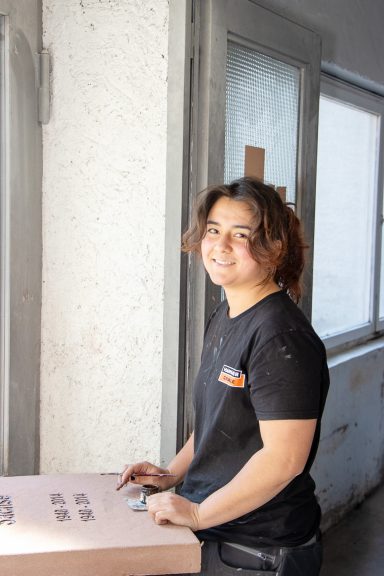Person mit lockigem Haar lächelt, während sie an einem Tisch steht. Licht fällt durch ein Fenster.