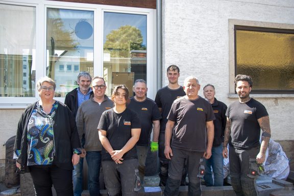 Gruppe von zehn Personen in Arbeitskleidung vor einem Gebäude, lächelnd und posierend.
