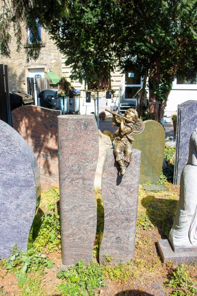 Grabsteine auf einem Friedhof, mit einem Engel, umgeben von Bäumen und Gebäuden im Hintergrund.