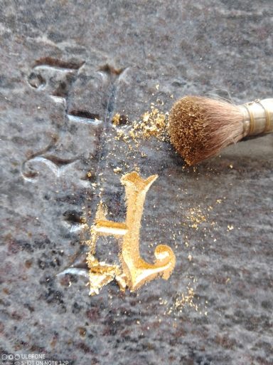 Goldene Initiale „H“ auf einer grauen Oberfläche, daneben ein Pinsel mit Goldstaub.