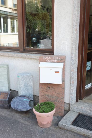 Weißer Briefkasten an einer Wand, daneben ein Topf mit grünen Pflanzen und Steinen.