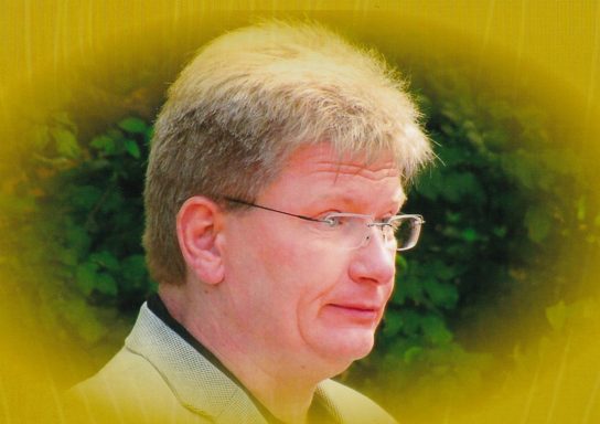 Mann mit kurzen blonden Haaren und Brille vor grünem Hintergrund.