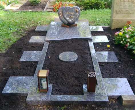 Gräber mit einem herzförmigen Grabstein und zwei Laternen, umgeben von Blumenbeeten.