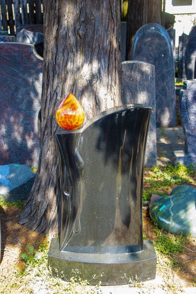 Schwarzer Grabstein neben einem Baum, mit einer leuchtenden orangefarbenen Kerze darauf.