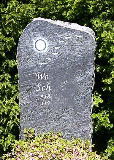 Grabstein aus grauem Stein mit eingravierter Schrift und Sonnenmotiv oben.