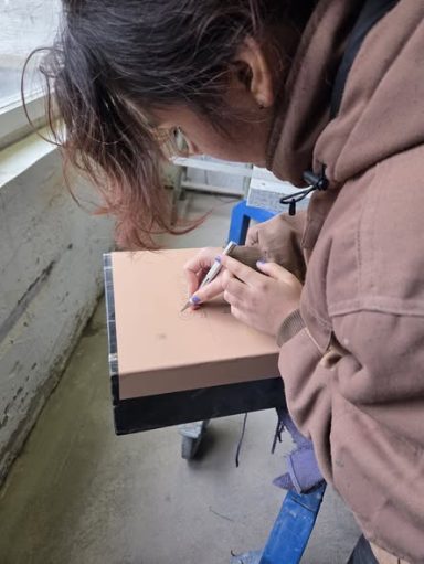 Eine Person zeichnet mit einem Stift auf eine braune Fläche.