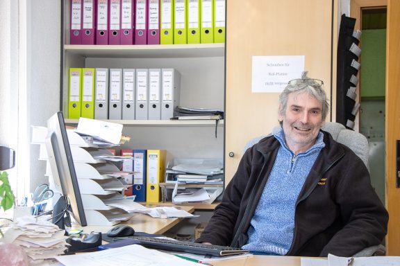 Lächelnder Mann am Schreibtisch mit Aktenordnern im Hintergrund.