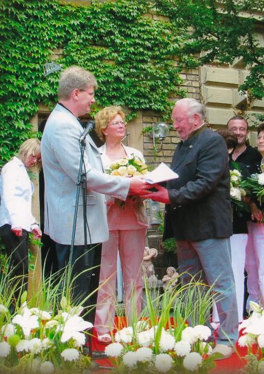 Zwei Männer und eine Frau überreichen sich Blumen bei einer Zeremonie im Freien.