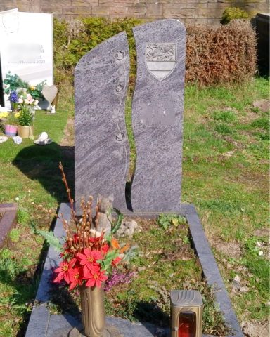 Dunkler, geschliffener Grabstein mit Blumenarrangement und Grünpflanzen im Hintergrund.