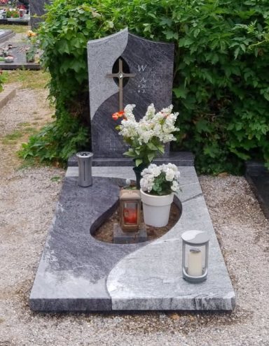 Grabstein mit Kreuz, Blumen und Kerzen, umgeben von Kies und grünen Sträuchern.