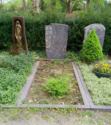 Drei Grabsteine in einer Gartenanlage mit Pflanzen und Blumen.
