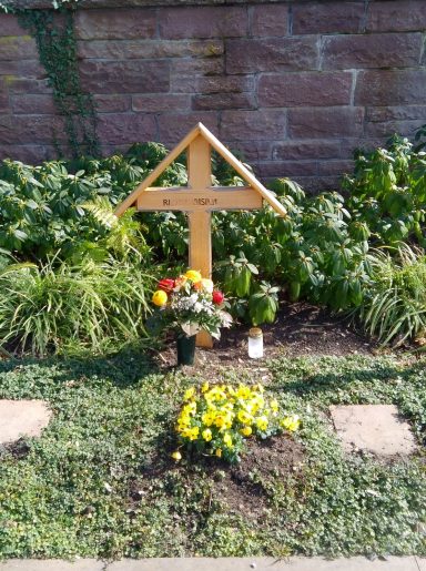 Holzkreuz mit Blumen und Kerze auf einer gepflegten Grabstätte.