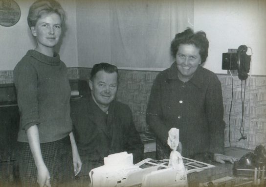 Drei Personen in einem Raum, eine Frau steht, zwei sitzen an einem Tisch mit einer Nähmaschine.