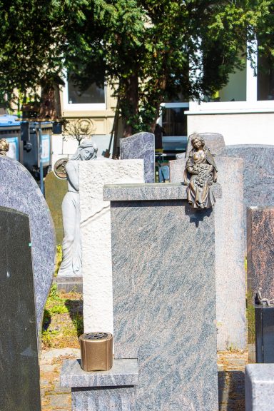 Gräber mit Grabsteinen und einer kleinen Statue auf einem Stein in einem Friedhofsbereich.